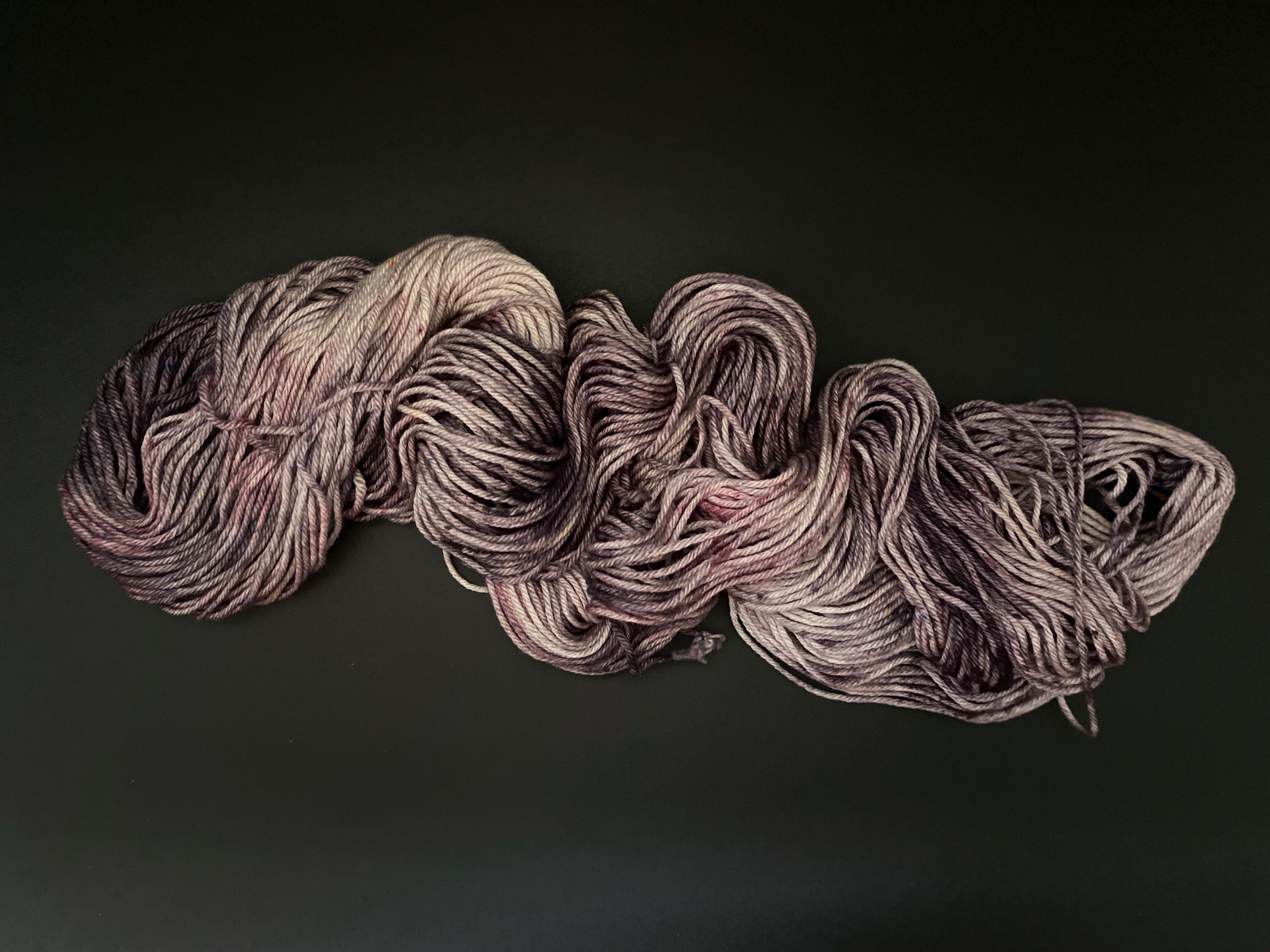 A single skein of hand dyed yarn, with various purple and mauve tones, draped on a black background.