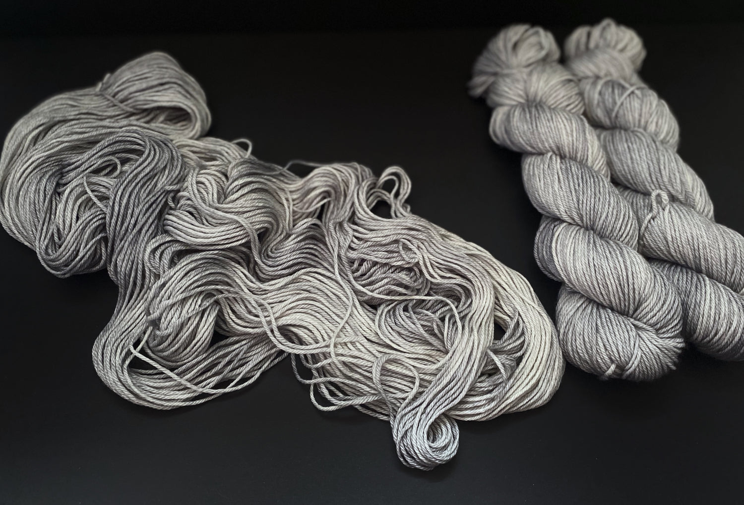 Three skeins of cool silver grey yarn, two on the left wound into hanks, and one draped flowingly on a black background.
