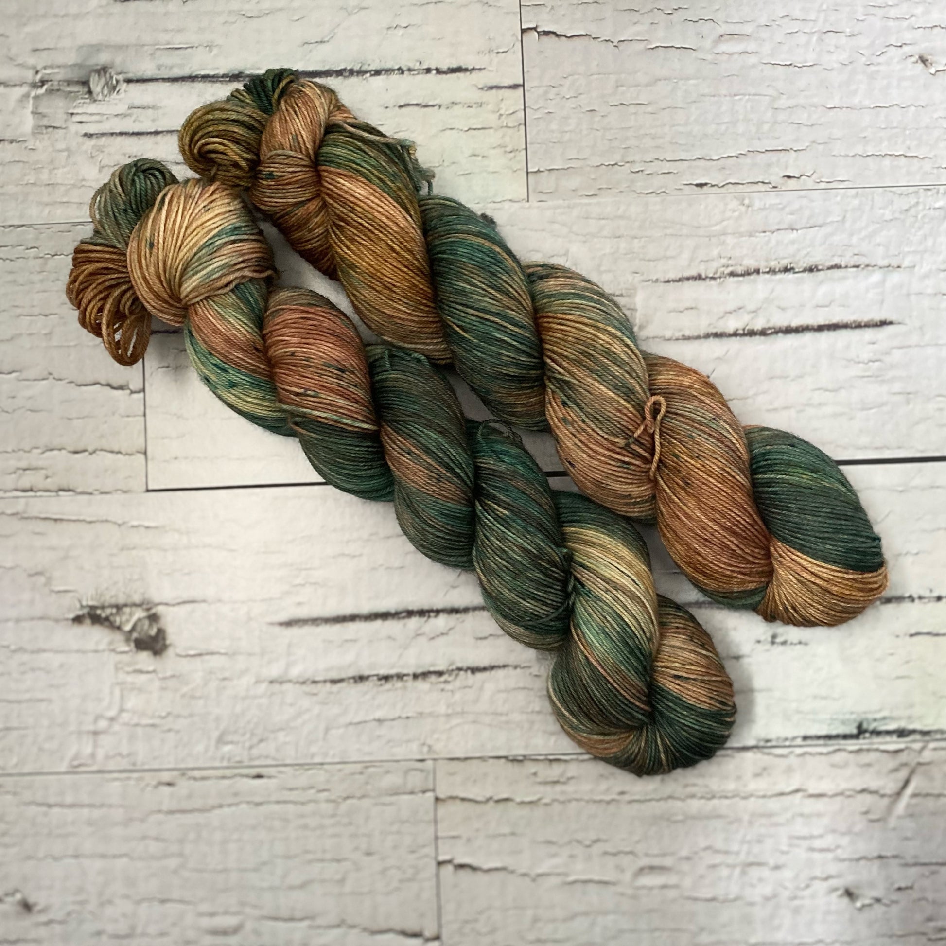 Two skeins of brown and green variegated hand dyed yarn laid on a white wood plank background.