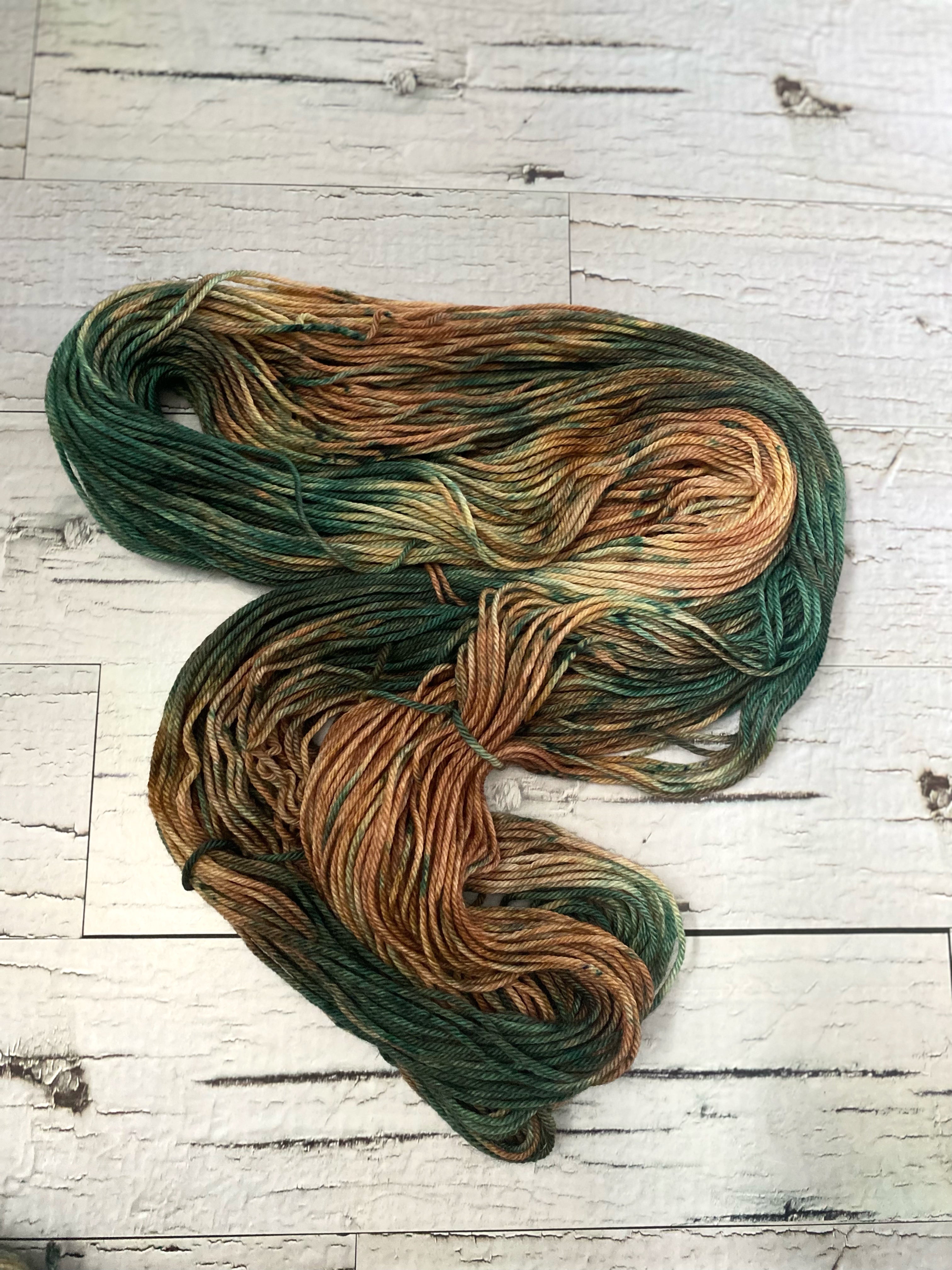 A hank of  variegated hand dyed yarn laid out on a white wood board background. The yarn consists of beautiful fawn brown and forest green tones.