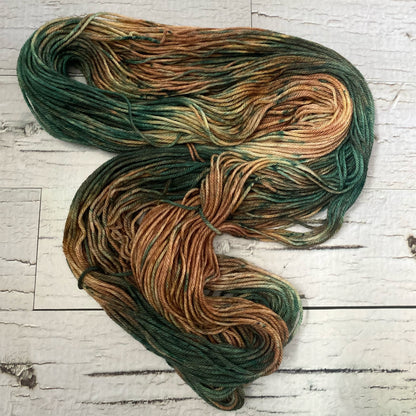 A hank of  variegated hand dyed yarn laid out on a white wood board background. The yarn consists of beautiful fawn brown and forest green tones.