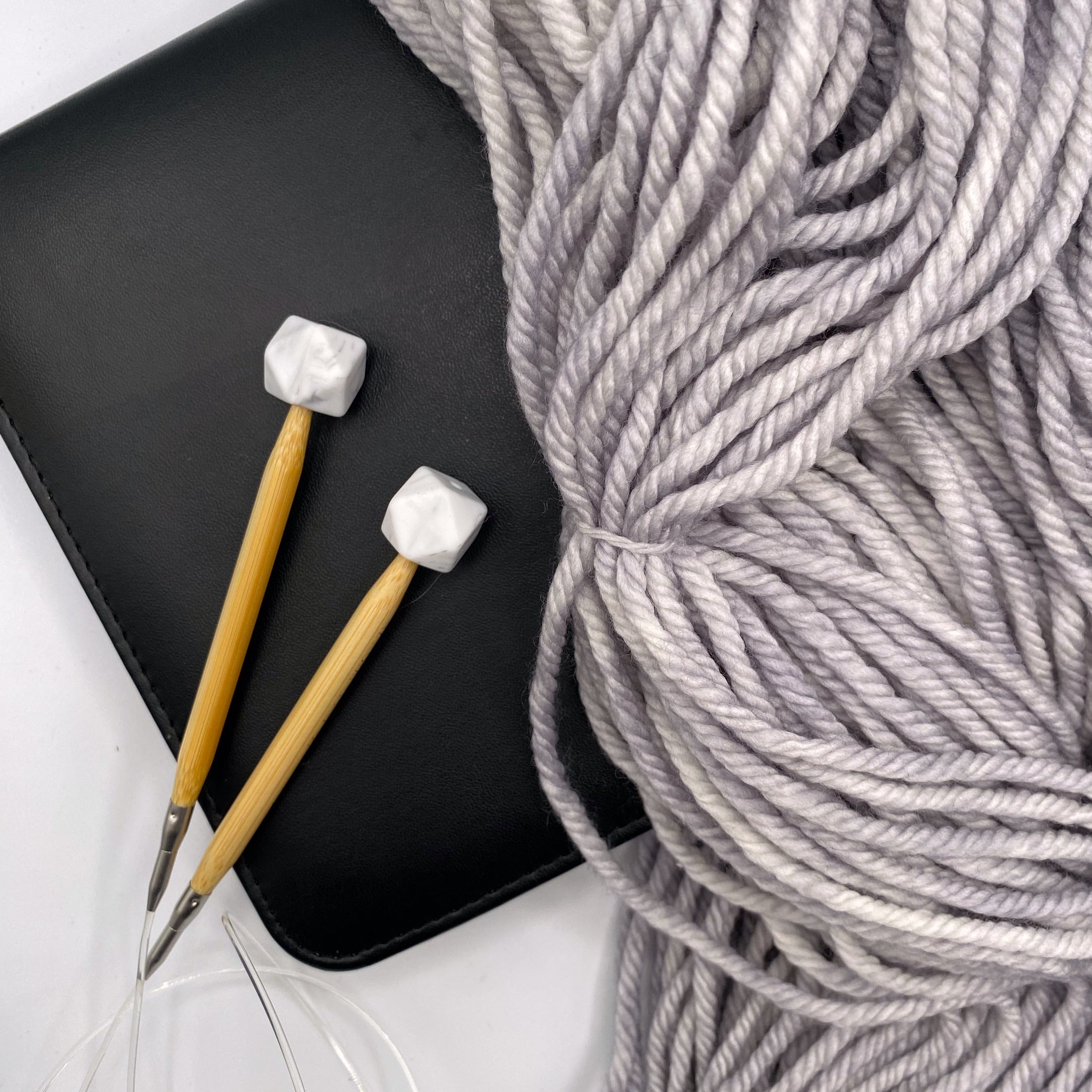 a set of knitting needles adorned with marble hexagon point protectors, laid on top of a faux leather needle case, beside our Everyday Bulky in Urban.