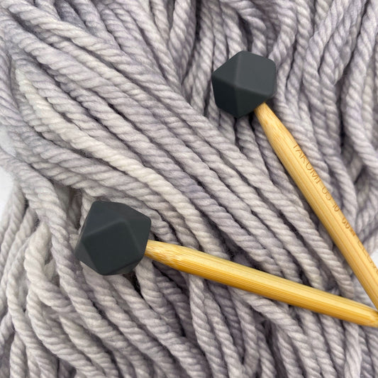 A set of knitting needles adorned with charcoal hexagon point protectors, laid on top of a spread of our Everyday Bulky in Urban.