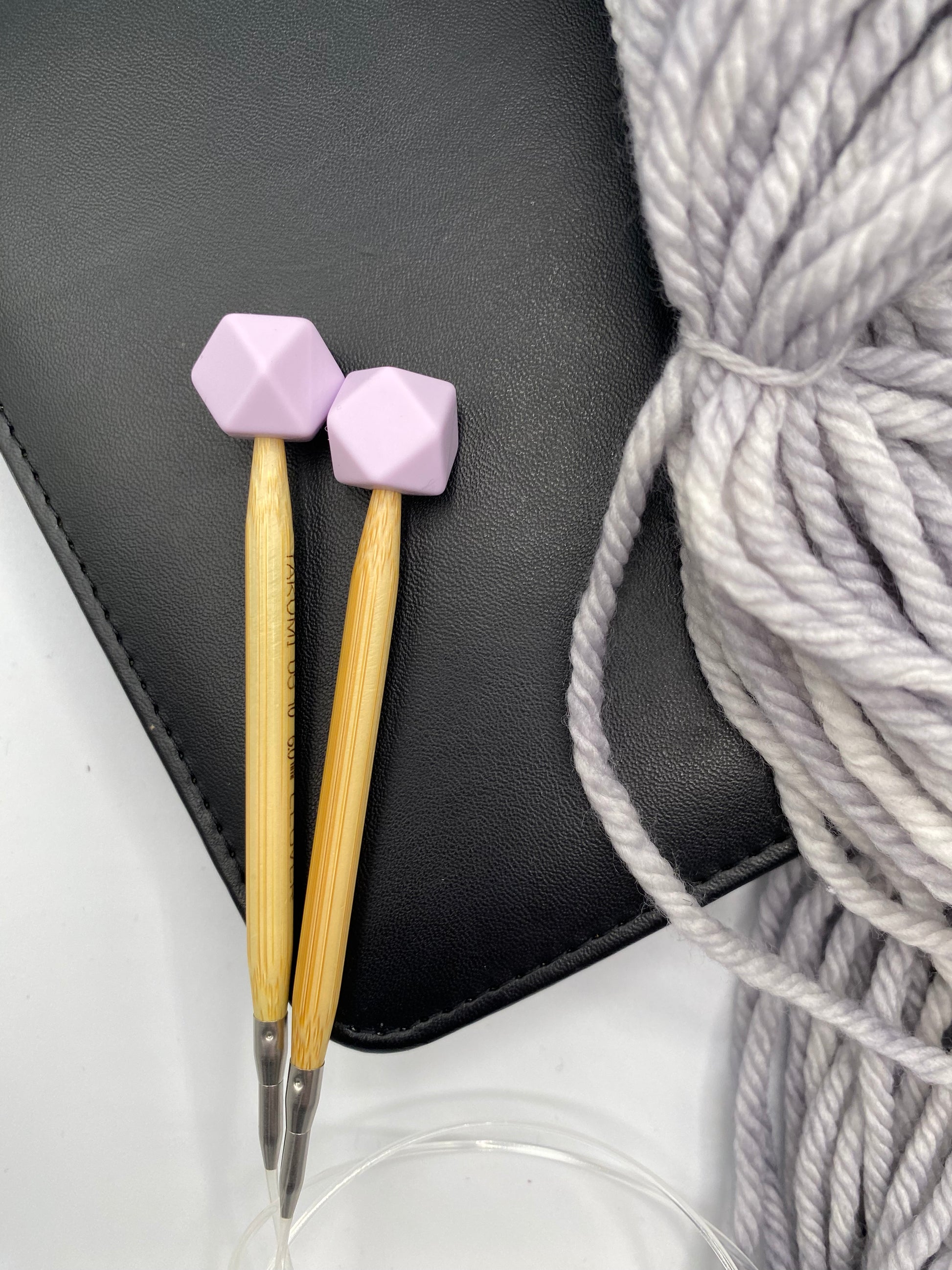 a set of knitting needles adorned with lilac hexagon point protectors, laid on top of a faux leather needle case, beside our Everyday Bulky in Urban.